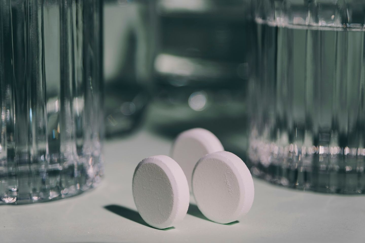 Tres comprimidos redondos y blancos sobre una mesa, entre dos vasos de agua desenfocados