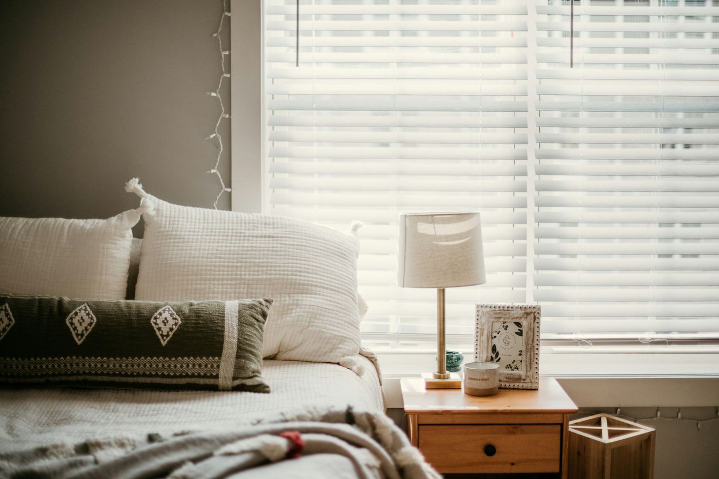 Dormitorio tranquilo con cojines blancos, una lámpara en la mesilla y luz natural que entra por la persiana
