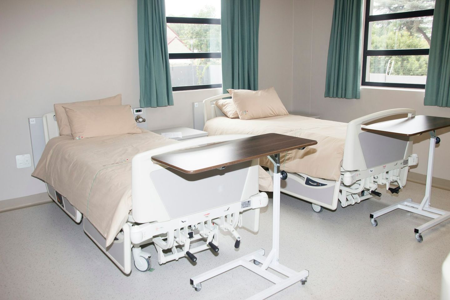 Habitación de hospital con dos camas, mesitas auxiliares y ventanas con cortinas verdes