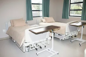 Habitación de hospital con dos camas, mesitas auxiliares y ventanas con cortinas verdes