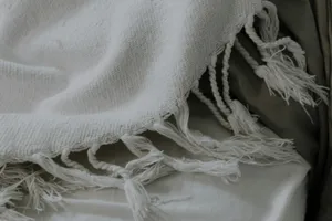 Manta blanca de tejido grueso con flecos, en primer plano, sobre una tela clara