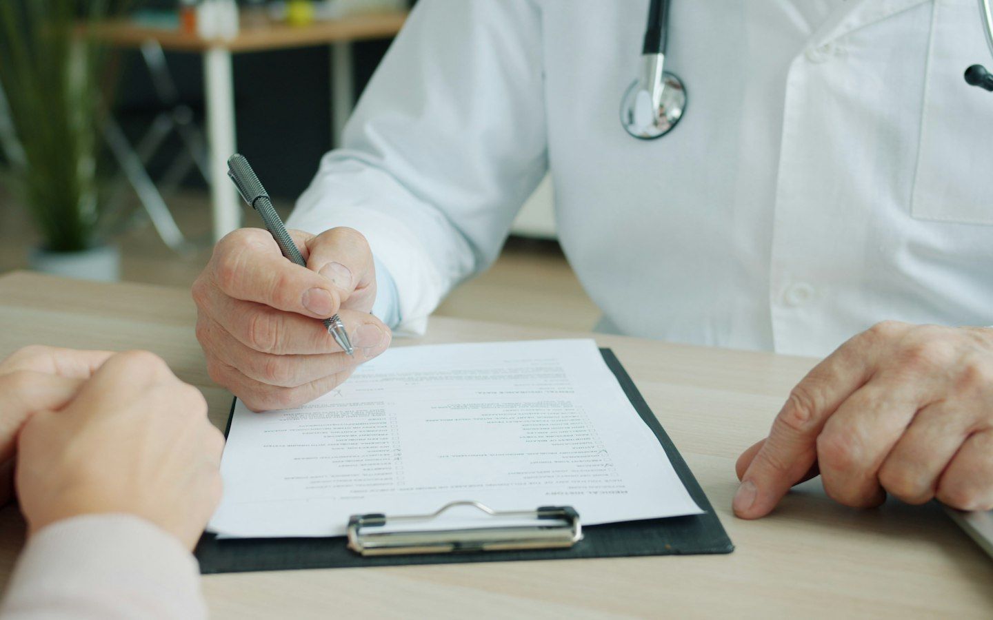 Manos de un médico con bata y estetoscopio que toma notas en una consulta, frente a las manos de un paciente