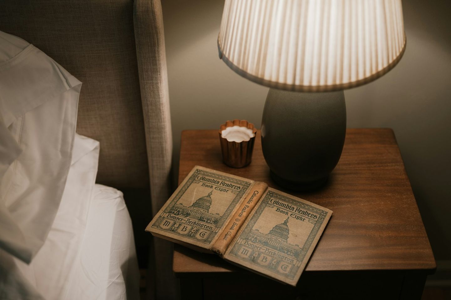 Mesilla de noche con una lámpara encendida, una vela pequeña y un libro antiguo junto a una cama