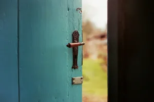 Puerta de madera azul turquesa entreabierta, con un picaporte antiguo y un jardín desenfocado al fondo